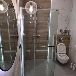 Nowoczesna łazienka z kabiną prysznicową typu walk-in, wykończona płytkami imitującymi drewno i marmur, oświetlona kulistymi lampami wiszącymi i ledowym paskiem wzdłuż ściany.