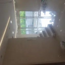 Wnętrze w trakcie prac wykończeniowych: ściany pokryte tynkiem dekoracyjnym, widoczne schody w surowym stanie, okno z widokiem na zieleń, oświetlenie sufitowe LED, puszki z farbami w tle.