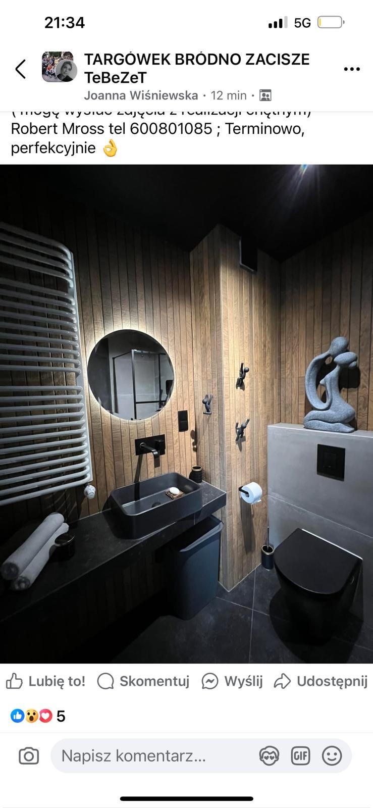 Nowoczesna łazienka z drewnianymi panelami ściennymi, czarną armaturą i okrągłym lustrem z podświetleniem LED. Dekoracyjna rzeźba na spłuczce dodaje elegancji.