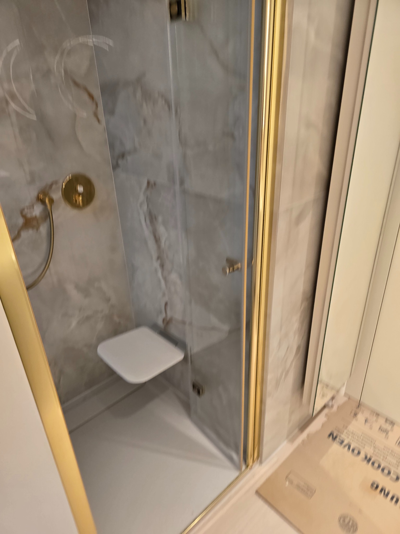 Nowoczesna kabina prysznicowa z marmurowymi płytkami, złotymi detalami i siedziskiem, widoczne zabezpieczenie podłogi kartonem.