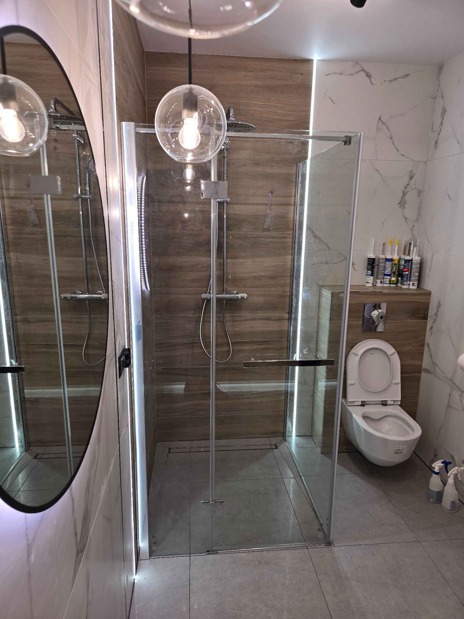 Nowoczesna łazienka z kabiną prysznicową typu walk-in, wykończona płytkami imitującymi drewno i marmur, oświetlona kulistymi lampami wiszącymi i ledowym paskiem wzdłuż ściany.