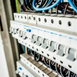 Szczegółowe ujęcie rozdzielnicy elektrycznej z widocznymi bezpiecznikami automatycznymi marki Hager i okablowaniem w kolorach niebieskim i czarnym, uchwycone pod lekkim kątem.