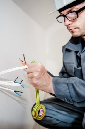 Mężczyzna w okularach i kombinezonie, klęczący przy ścianie, obcina przewody elektryczne za pomocą kombinerek; widoczna taśma izolacyjna.