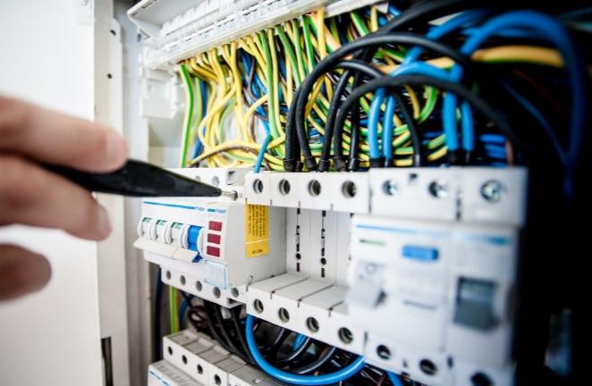 Próba napięcia w skrzynce rozdzielczej z bezpiecznikami, widoczne kolorowe przewody i ręka z próbnikiem.