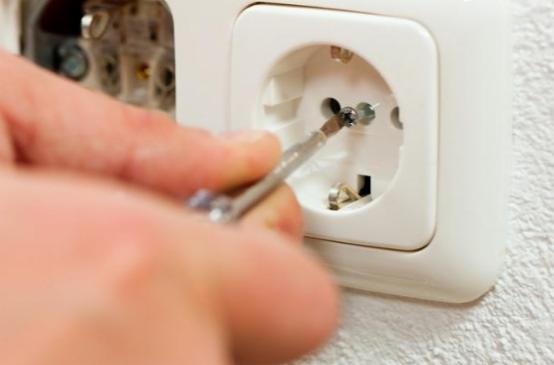 Dłoń dokręca śrubokrętem śrubę w gniazdku elektrycznym, widoczna puszka instalacyjna w ścianie.