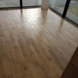 Ułożona podłoga z płytek imitujących drewno, widok z niskiej perspektywy, duże okna z czarnymi ramami w tle.