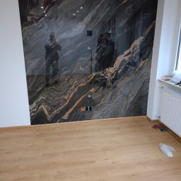 Daniel Kuciński - Ściana wykończona płytami imitującymi marmur z widocznymi gniazdkami elektrycznymi, na podłodze panele. Widoczne odbicia osób na ścianie.