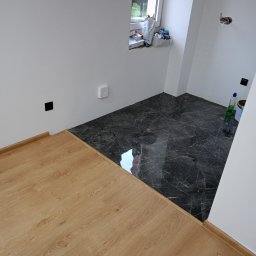 Daniel Kuciński - Połączenie dwóch rodzajów podłóg: ciemne płytki imitujące marmur i jasne panele drewnopodobne. Widoczne gniazdka elektryczne i okno z akcesoriami.