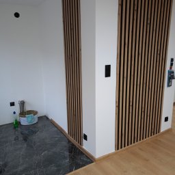 Daniel Kuciński - Nowoczesne wykończenie wnętrza: białe ściany z drewnianymi lamelami, marmurowe płytki podłogowe przechodzące w panele. Widoczne gniazdka elektryczne i akcesoria malarskie.