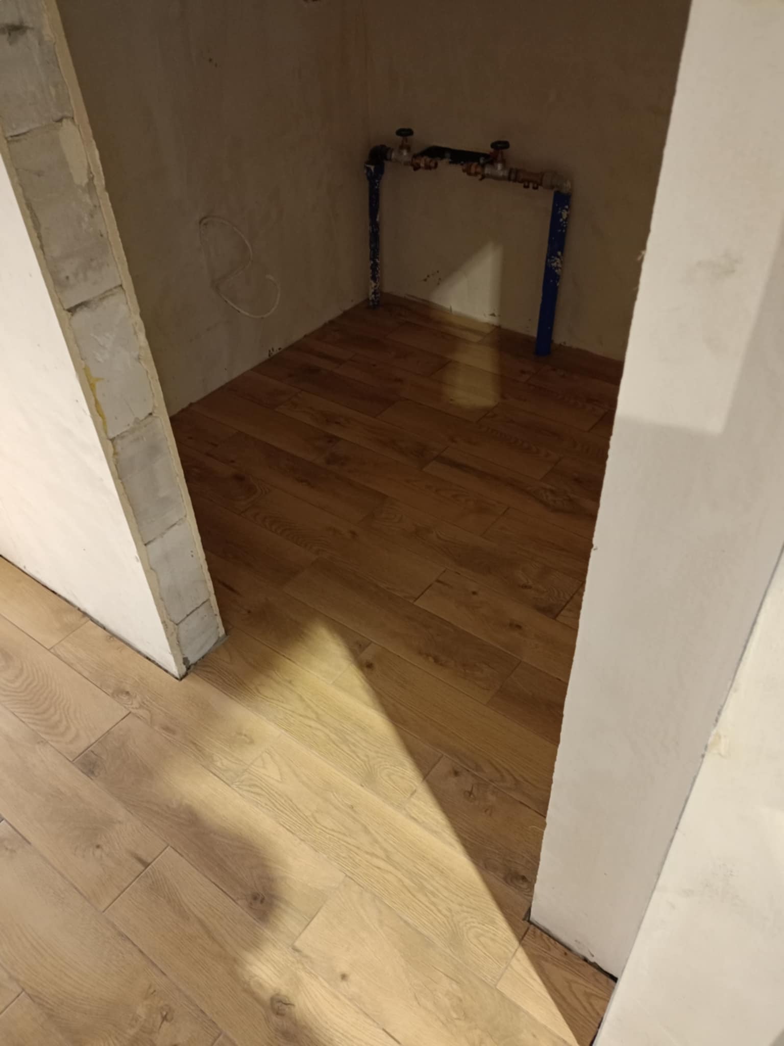 Małe pomieszczenie z drewnianą podłogą ułożoną w jodełkę, nieotynkowane ściany i niebieskie rury z zaworami przy podłodze.