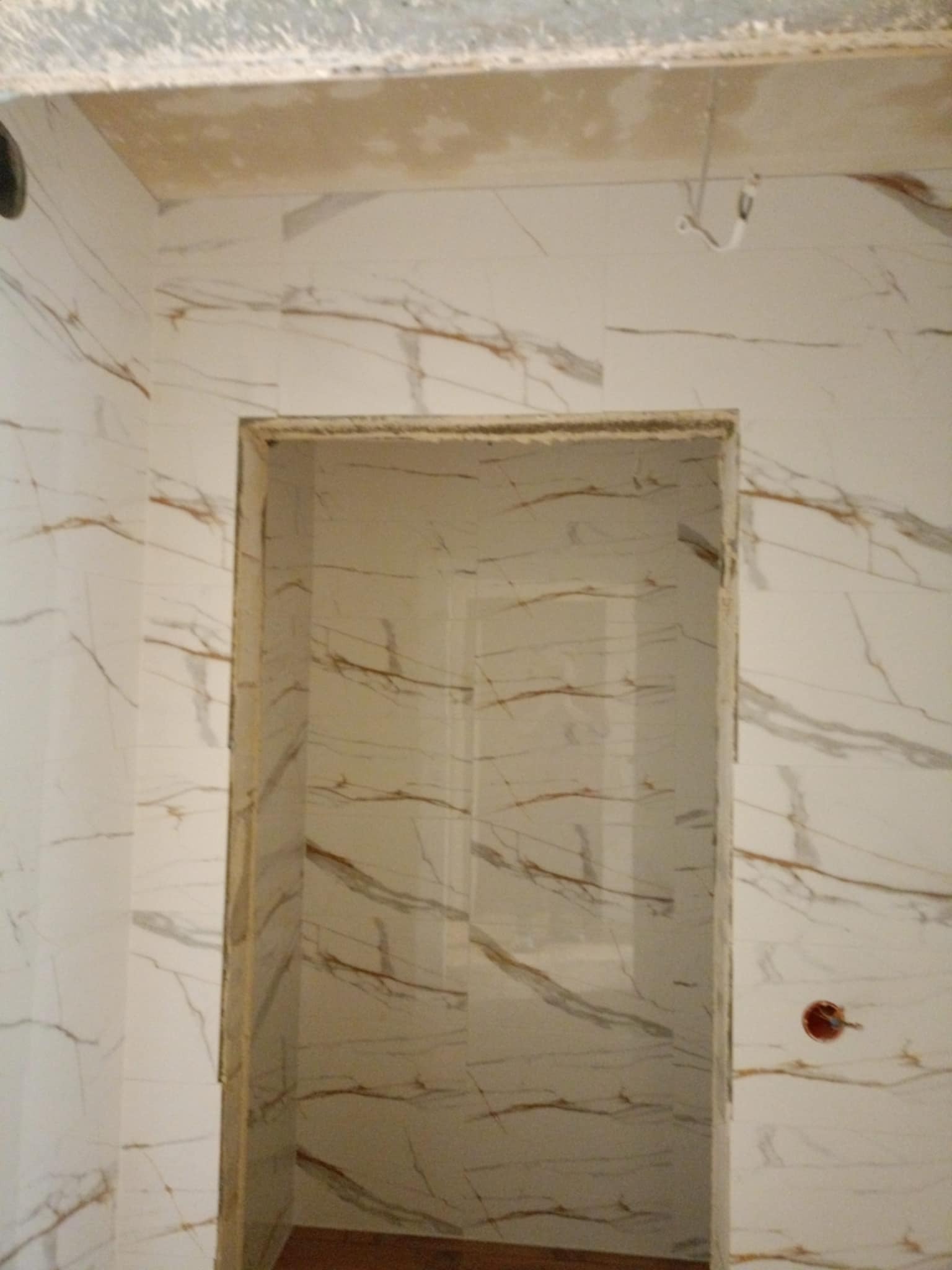 Ściany i framuga drzwi wyłożone białymi płytkami imitującymi marmur z brązowymi żyłami, widoczne niedokończone prace sufitowe i instalacja elektryczna w puszce.