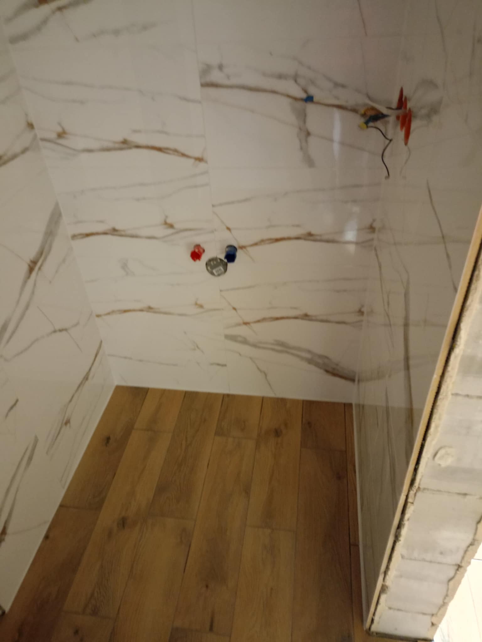 Wnętrze kabiny prysznicowej w trakcie wykańczania: marmurowe płytki ścienne z brązowymi żyłkami, drewnopodobna podłoga, wystające przewody elektryczne i rury wodne.