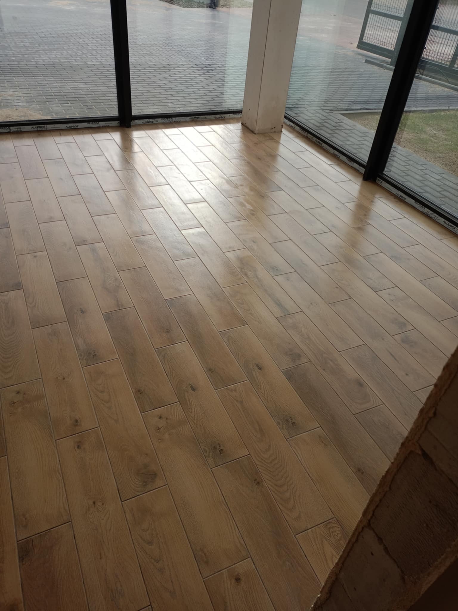 Ułożona podłoga z płytek imitujących drewno, widok z niskiej perspektywy, duże okna z czarnymi ramami w tle.