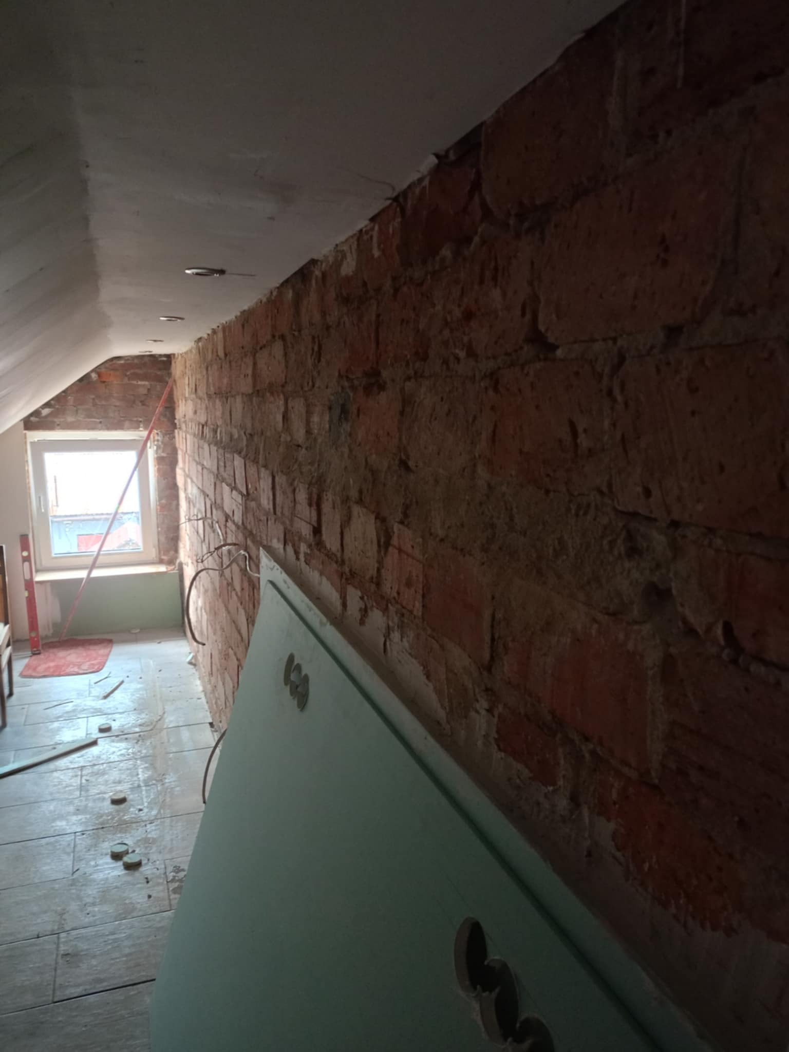 Ściana z czerwonej cegły w trakcie remontu poddasza, widoczne przewody elektryczne, płyta karton-gips oparta o ścianę, okno w szczycie dachu, poziomica i fragment podłogi z płytek.