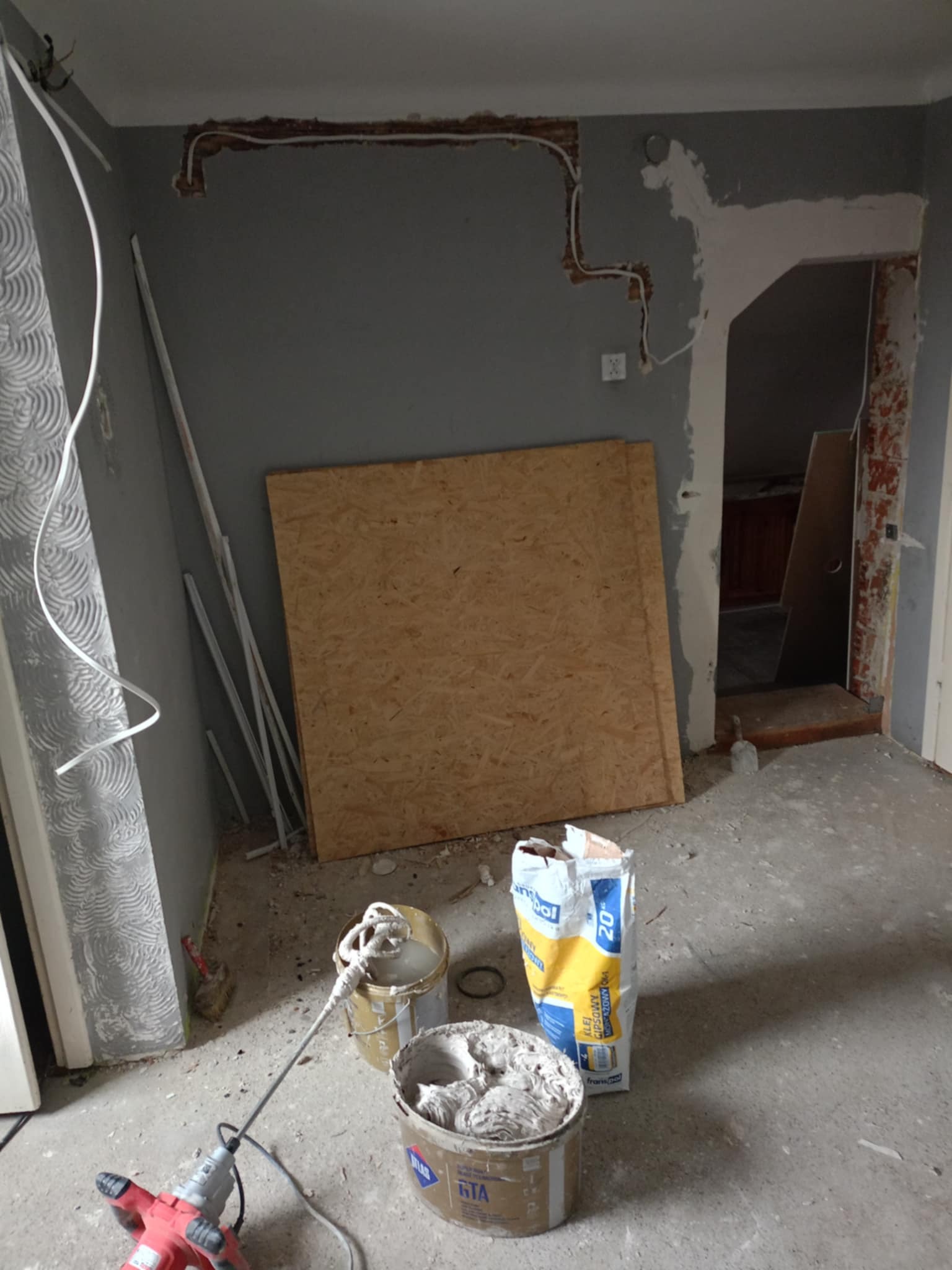 Wnętrze w trakcie remontu: widoczne ślady po instalacji elektrycznej w ścianie, płyta OSB oparta o ścianę, wiadro z zaprawą, mieszadło elektryczne i worek z klejem gipsowym.