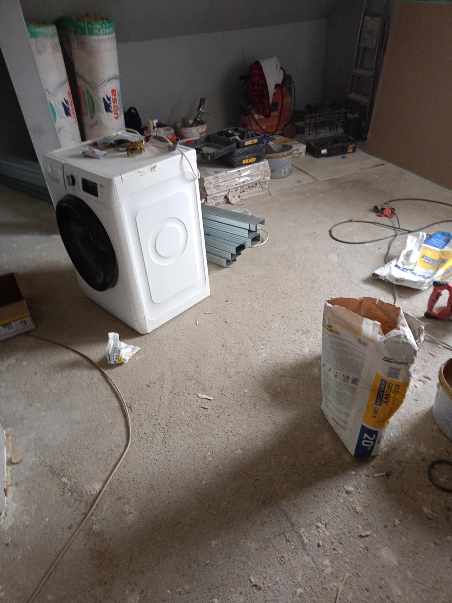 Pralka w surowym stanie pomieszczenia podczas prac remontowych, obok narzędzia, profile metalowe i worek z klejem gipsowym.