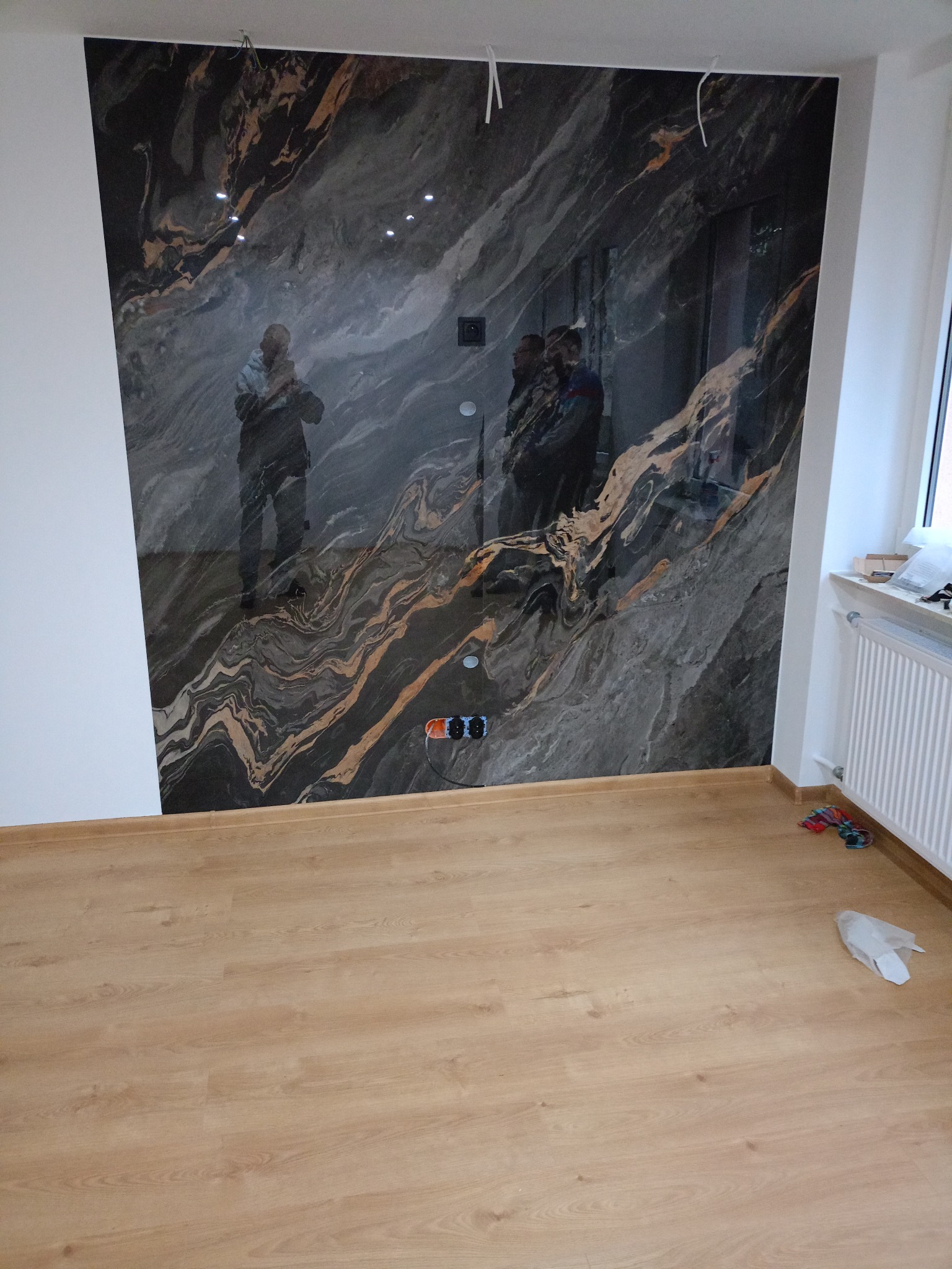 Ściana wykończona płytami imitującymi marmur z widocznymi gniazdkami elektrycznymi, na podłodze panele. Widoczne odbicia osób na ścianie.