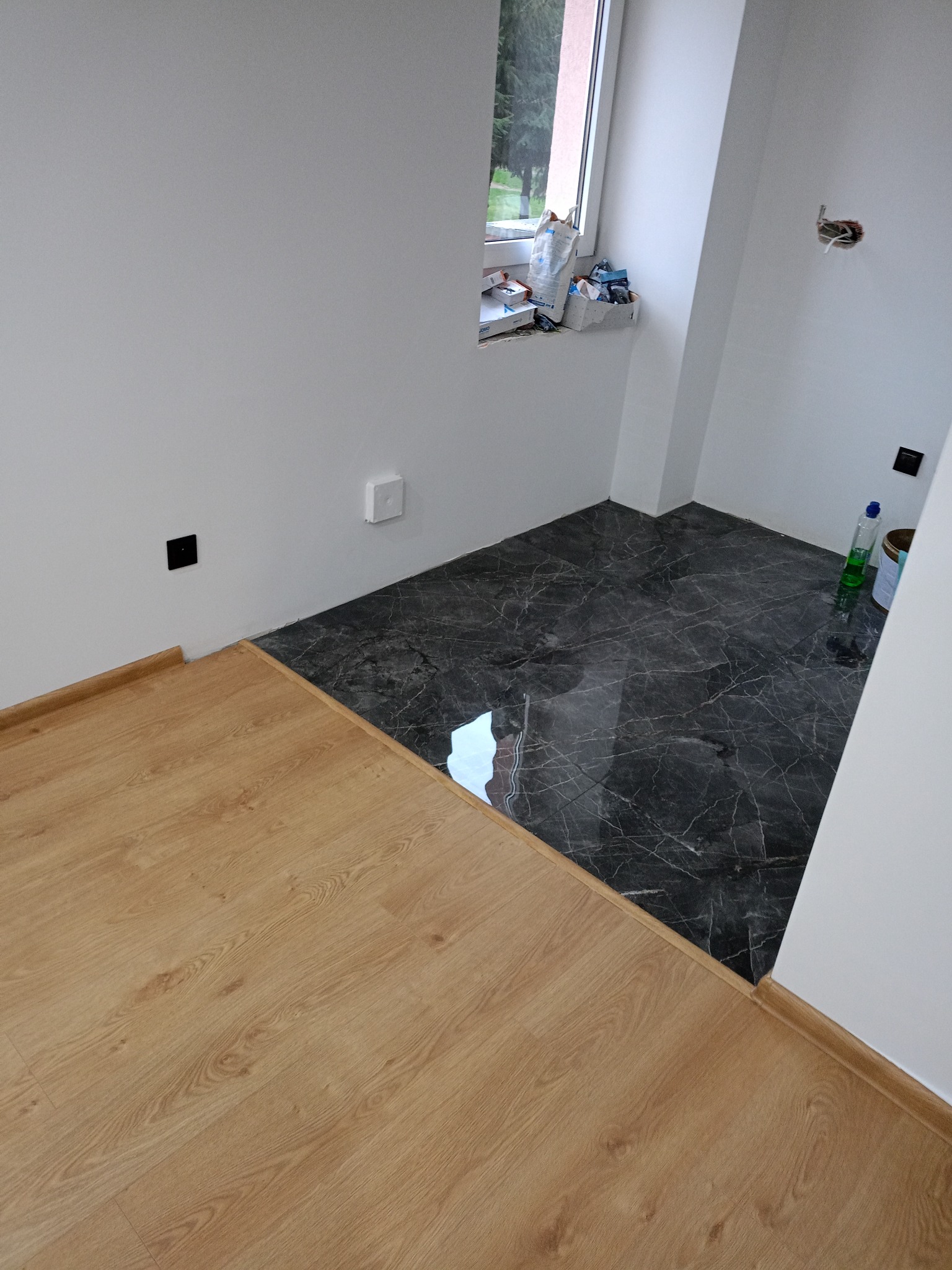 Połączenie dwóch rodzajów podłóg: ciemne płytki imitujące marmur i jasne panele drewnopodobne. Widoczne gniazdka elektryczne i okno z akcesoriami.