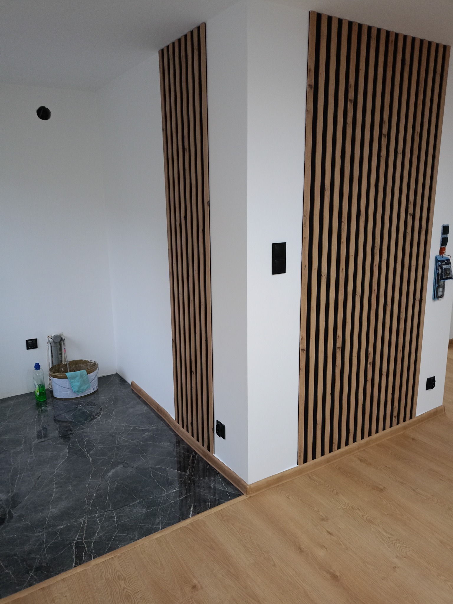 Nowoczesne wykończenie wnętrza: białe ściany z drewnianymi lamelami, marmurowe płytki podłogowe przechodzące w panele. Widoczne gniazdka elektryczne i akcesoria malarskie.