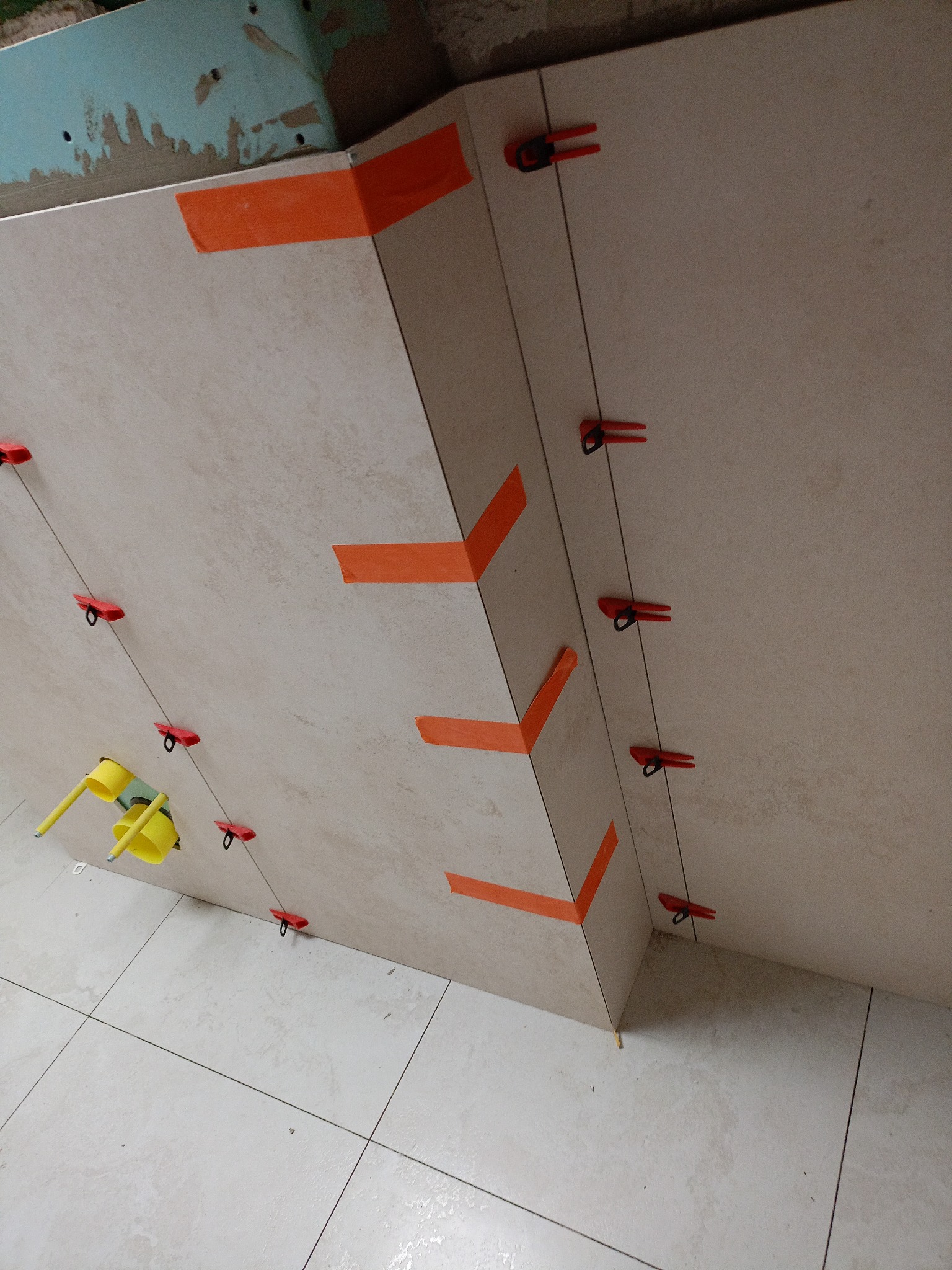 Układanie beżowych płytek na ścianie z użyciem pomarańczowej taśmy i czerwonych klipsów poziomujących. Widoczne narzędzie do poziomowania płytek.