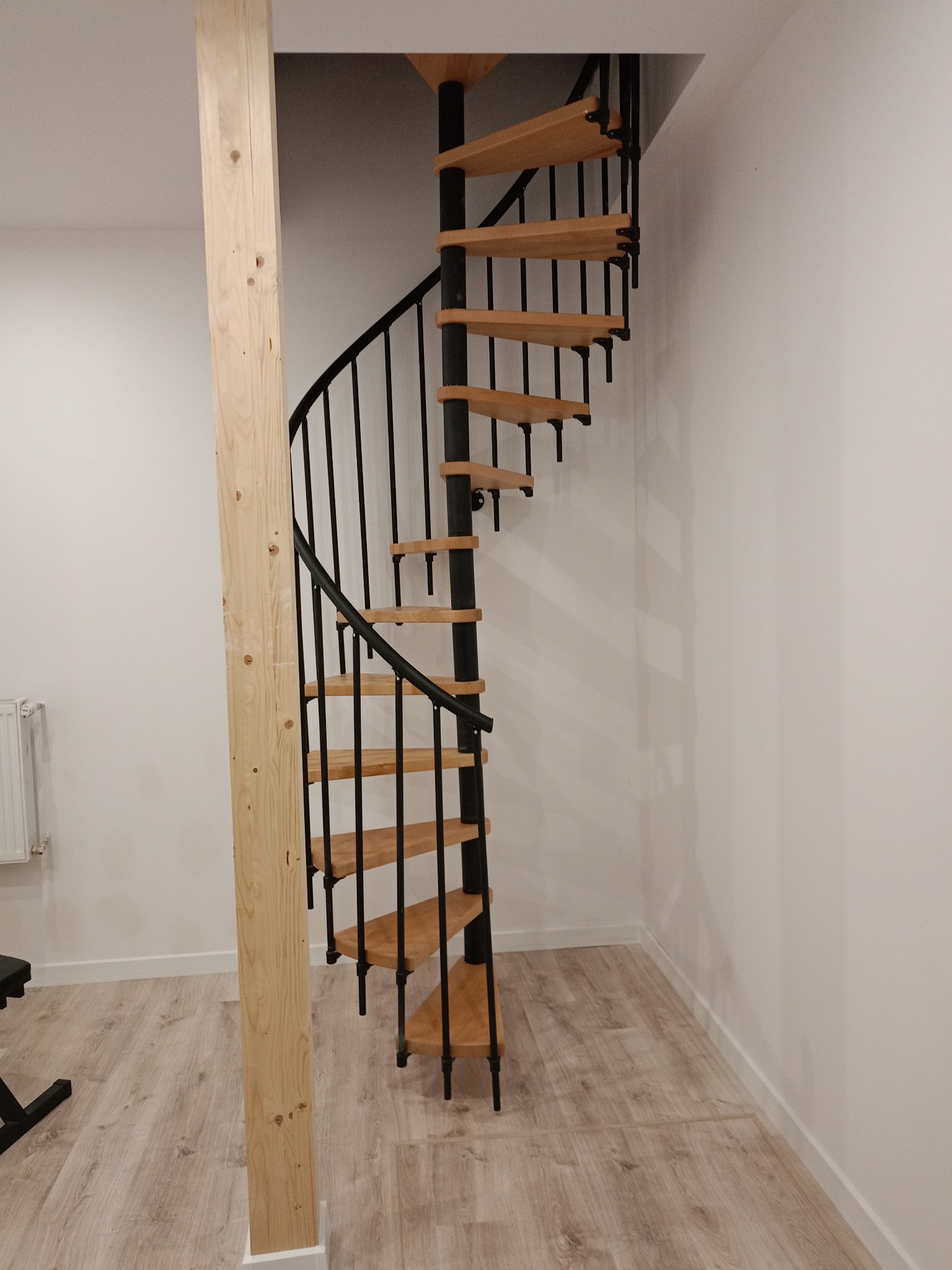 Spiralne schody z drewnianymi stopniami i czarną metalową konstrukcją, umieszczone w rogu pomieszczenia z jasnymi ścianami i drewnopodobną podłogą.
