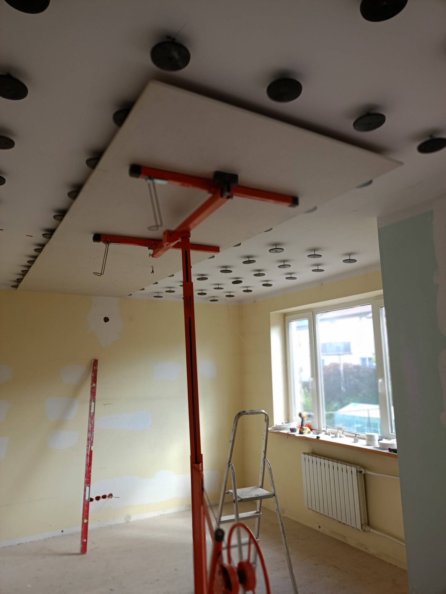 Montaż płyty gipsowej na suficie za pomocą podnośnika. Widoczne profile i narzędzia. Ściany przygotowane do malowania. Okno z parapetem.
