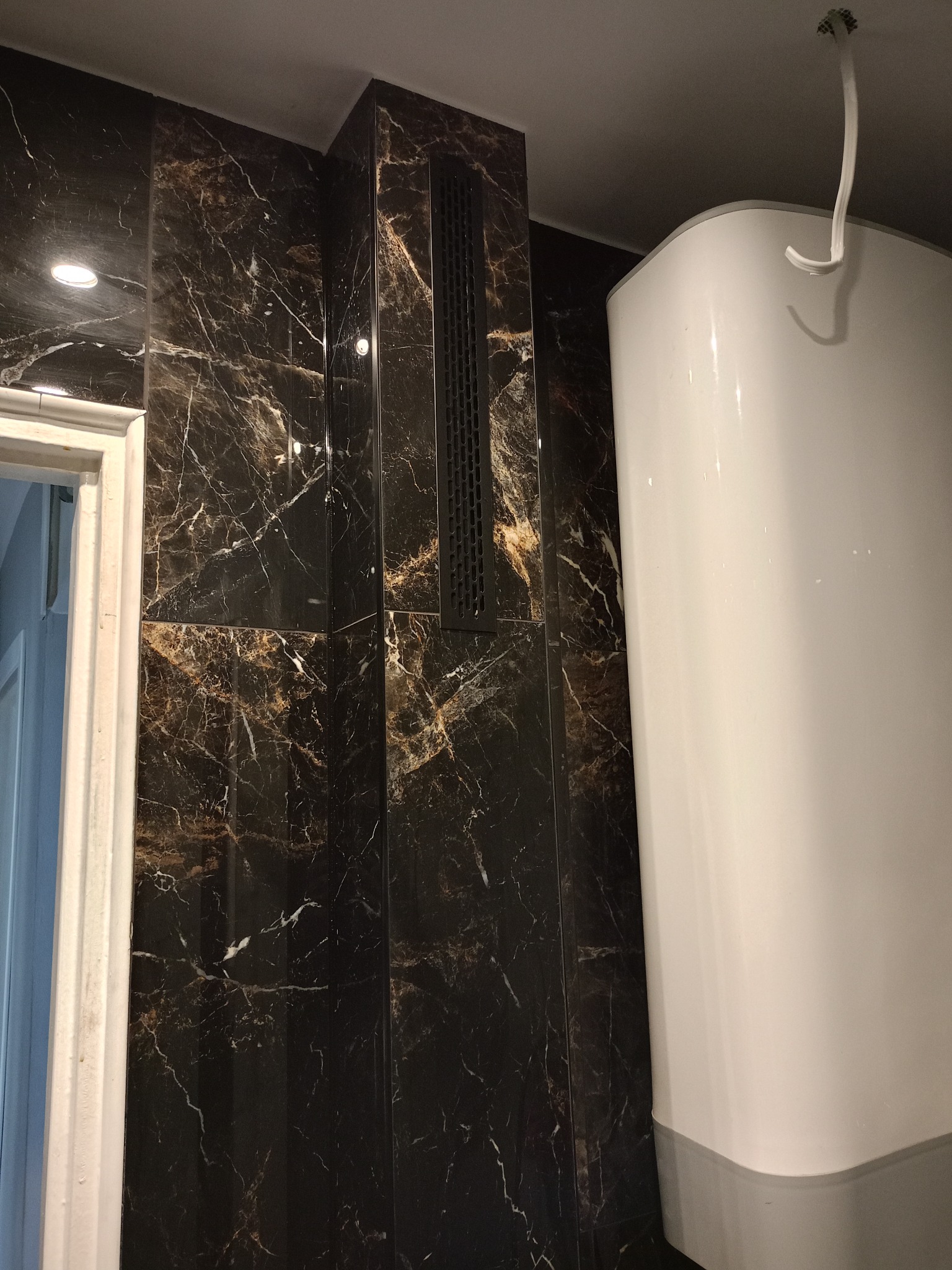 Wykończona łazienka z czarnymi płytkami imitującymi marmur, widoczny bojler i kratka wentylacyjna. Fragment ościeżnicy drzwi po lewej stronie kadru.