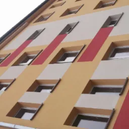 Wielopiętrowy budynek z pomalowaną elewacją w geometryczne wzory: żółty, biały i czerwony, okna z wystającymi parapetami.