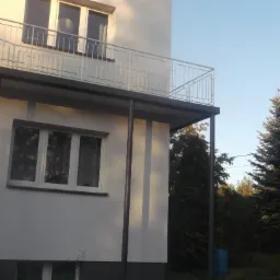 Boczny widok na dwukondygnacyjny dom z białymi ścianami, szarym cokołem i balkonem z metalową balustradą, wspartym na metalowych słupach, otoczony zielenią.