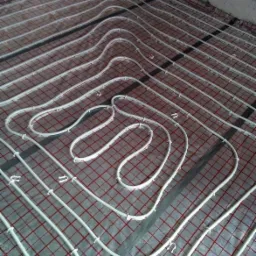 Rozłożona instalacja ogrzewania podłogowego na folii z siatką, rurki przymocowane plastikowymi uchwytami, widoczne fragmenty ściany.