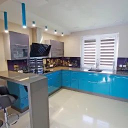 Nowoczesna kuchnia z błękitnymi szafkami, szarym blatem i panelem szklanym z motywem nocnej panoramy miasta. Widoczne krzesła barowe i oświetlenie punktowe.