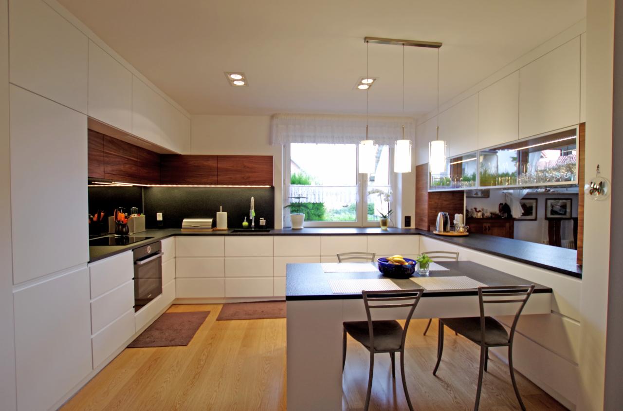 Nowoczesna kuchnia z białymi szafkami, drewnianymi akcentami i czarnym blatem, z wyspą i krzesłami barowymi, oświetlona wiszącymi lampami.