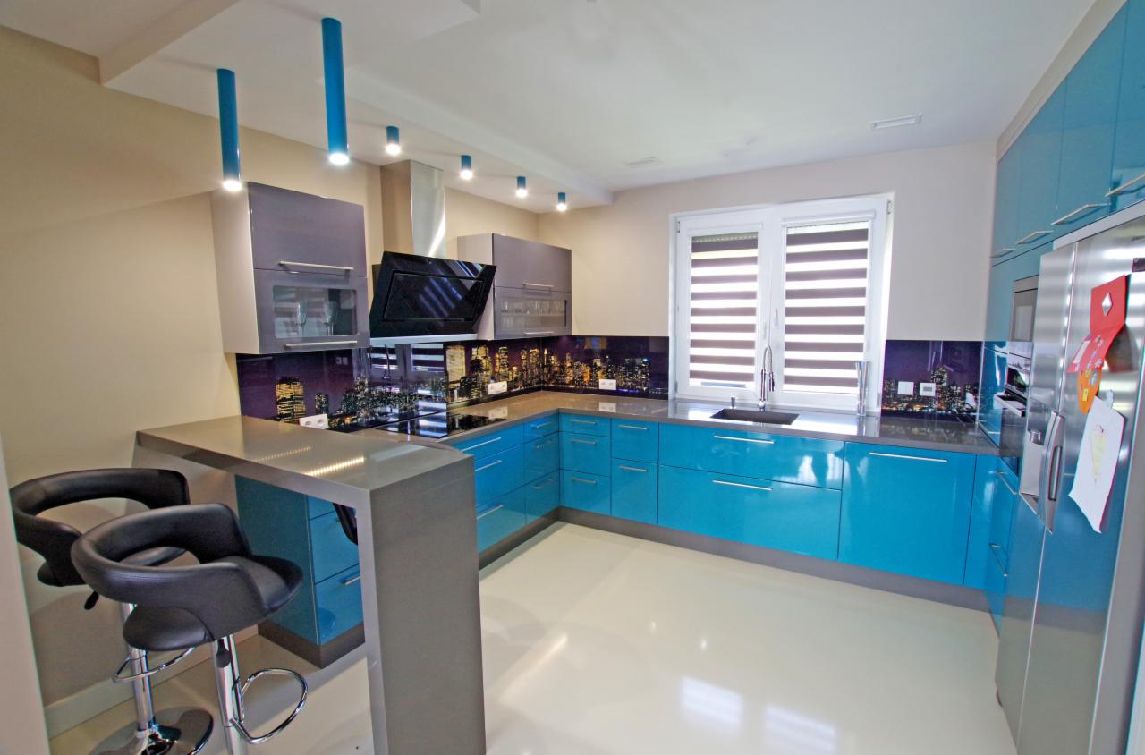 Nowoczesna kuchnia z błękitnymi szafkami, szarym blatem i panelem szklanym z motywem nocnej panoramy miasta. Widoczne krzesła barowe i oświetlenie punktowe.