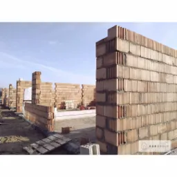 Wznoszenie ścian domu z bloczków ceramicznych Porotherm, widoczne spoiny i pustaki w pionowym przekroju, fundamenty w tle, logo firmy w prawym dolnym rogu.