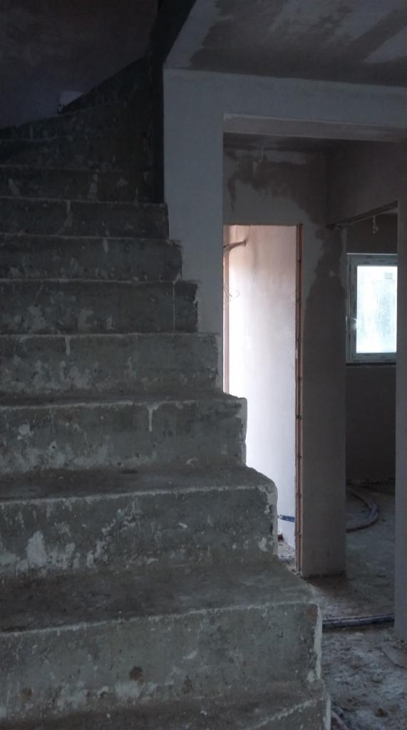 Surowe betonowe schody wewnętrzne w trakcie budowy lub remontu, widoczne ślady po szalunku, w tle fragment ściany i okno.