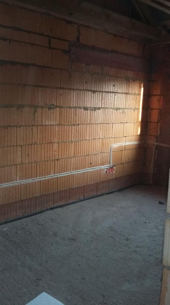 Wnętrze surowego domu w trakcie budowy: ściany z bloczków ceramicznych, betonowa posadzka, widoczne przewody elektryczne w peszlach. Promień słoneczny przebija przez otwór okienny.