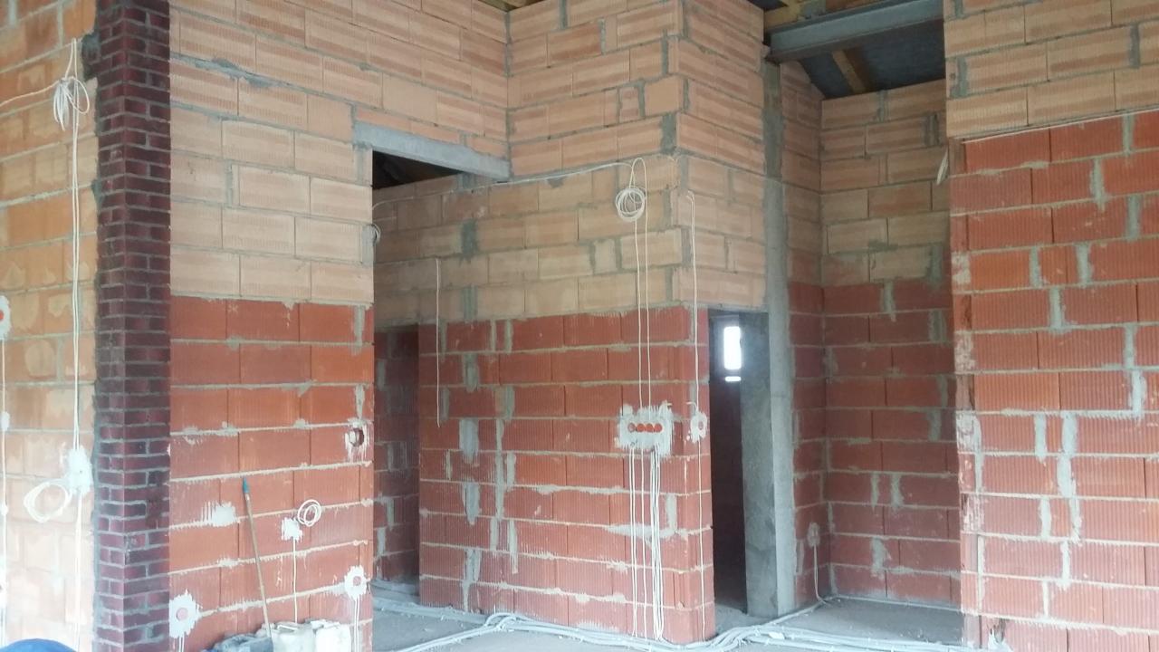 Wnętrze domu w trakcie budowy: ściany z czerwonej cegły, widoczne przewody elektryczne, surowy stan wykończenia.