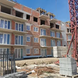 Wielopiętrowy budynek w trakcie wznoszenia, widoczne surowe ściany z czerwonej cegły, zamontowane okna z folią ochronną, wystające płyty balkonowe i czerwony żuraw budowlany na tle błękitnego nieba.