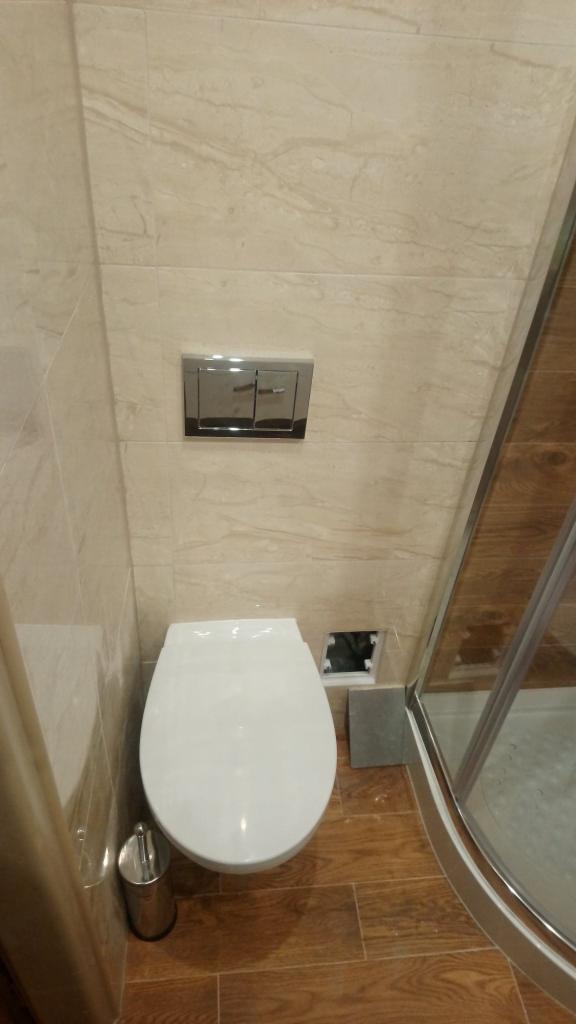 Nowoczesna toaleta podwieszana z chromowanym przyciskiem spłukującym i szczotką toaletową, obok kabiny prysznicowej z drewnopodobną podłogą i beżowymi płytkami ściennymi.