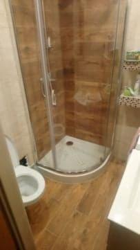 Narożna kabina prysznicowa z przezroczystymi drzwiami, biały brodzik i toaleta w małej łazience wykończonej płytkami imitującymi drewno, widok z góry.