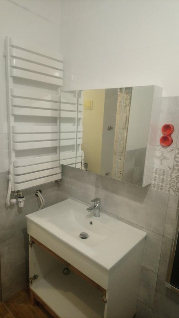 Nowoczesna łazienka z umywalką w szafce z otwartymi drzwiczkami, grzejnikiem drabinkowym i lustrem z szafką, szare płytki na ścianach z dekoracyjnym wzorem w rogu.