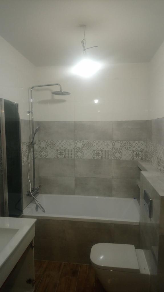 Wykończona łazienka z wanną obudowaną brązowymi płytkami, nad nią szare płytki z dekoracyjnym pasem, kabina prysznicowa po lewej, umywalka z szafką i wisząca toaleta, drewnopodobne płytki...