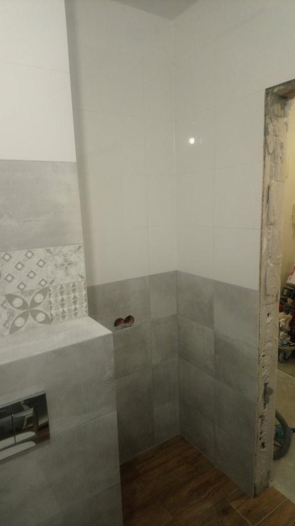 Świeżo wykafelkowana ściana łazienki z szarymi i białymi płytkami, widoczne gniazdka elektryczne i fragment drewnopodobnej podłogi, krawędź drzwi w stanie surowym.