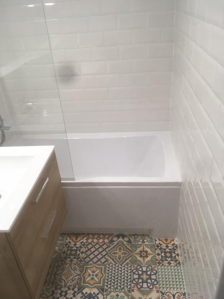 Biała wanna z szklanym parawanem, umywalka z drewnianą szafką i podłoga wyłożona wzorzystymi, kolorowymi płytkami w łazience po remoncie.