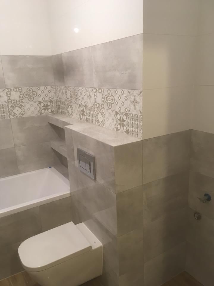 Wykończona łazienka w szarościach z dekoracyjnym pasem płytek, zabudowana wanna z półkami i podwieszana toaleta z chromowanym przyciskiem spłukującym.