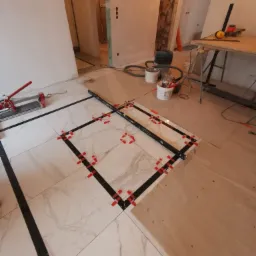 Układanie białych płytek imitujących marmur z czarnymi paskami dekoracyjnymi, używając poziomicy i czerwonych klipsów poziomujących, w trakcie prac wykończeniowych w pomieszczeniu.