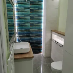 Nowoczesna łazienka z kabiną prysznicową wyłożoną płytkami w odcieniach błękitu i brązu, umywalką nablatową na drewnianym blacie, grzejnikiem drabinkowym i szarą podłogą.