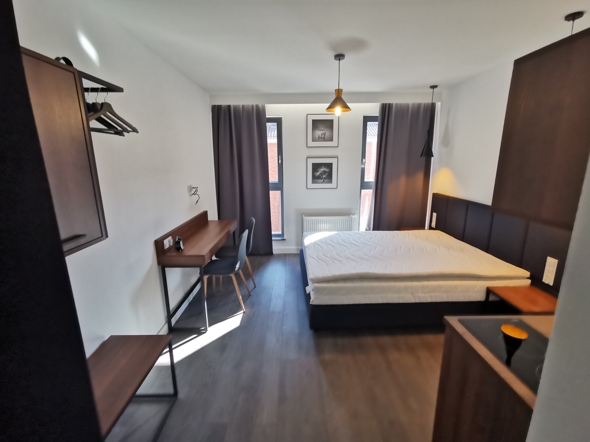 Nowocześnie urządzone studio z łóżkiem, biurkiem, krzesłami, szafą i aneksem kuchennym, oświetlone wiszącymi lampami i naturalnym światłem wpadającym przez okna z szarymi zasłonami.