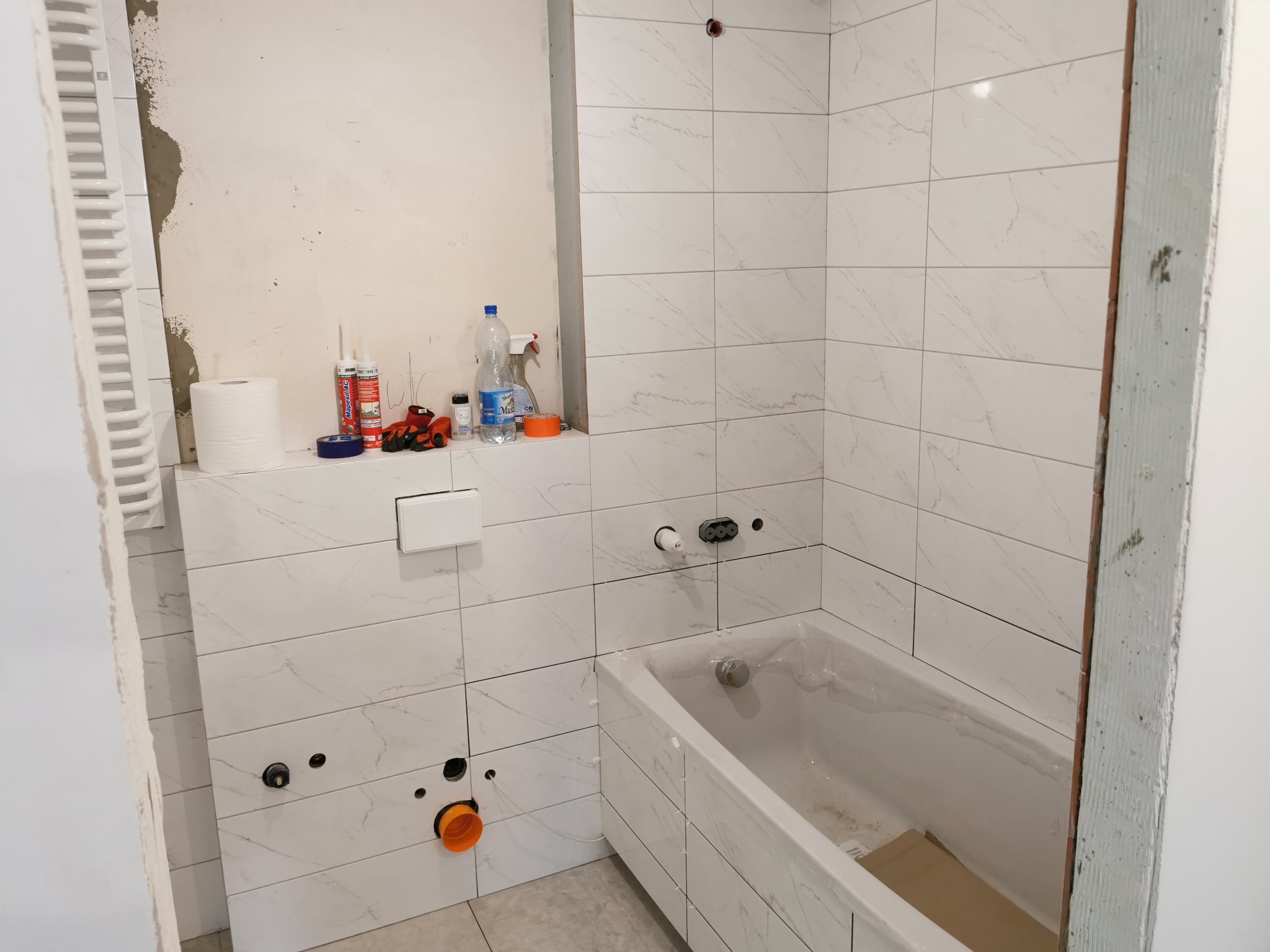 Wnętrze łazienki w trakcie prac: białe płytki imitujące marmur na ścianach, częściowo wykończone, widoczne przygotowane otwory na armaturę, wanna zabezpieczona folią, obok ściana z surowym...