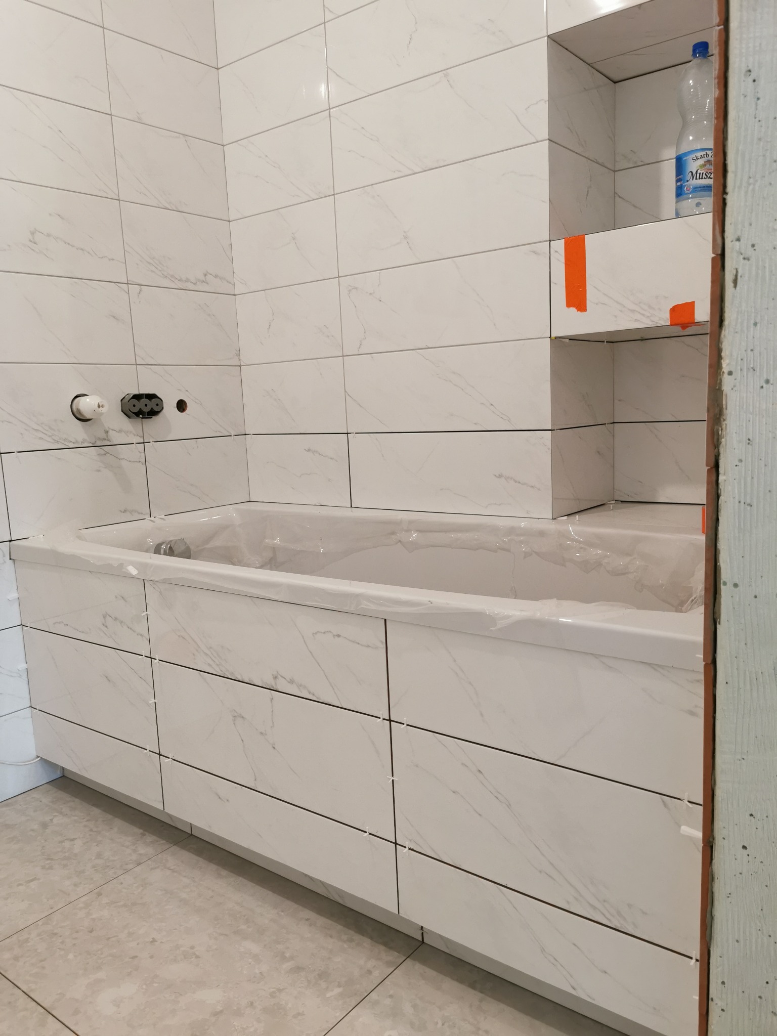 Biała wanna obudowana płytkami imitującymi marmur, z widocznymi krzyżykami dystansowymi, półka w ścianie wyłożona tymi samymi płytkami, zabezpieczona folią przed zabrudzeniem, butelka płynu...